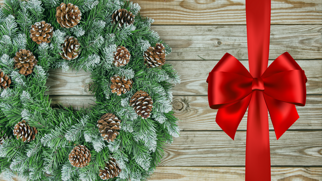 An evergreen wreath with pinecones sitting on a wooden table with a red ribbon bow on the side.