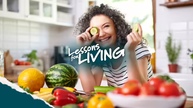 A woman holding a slice of cucumber over her eye with lots of fruits and vegetables in front of her in a kitchen.