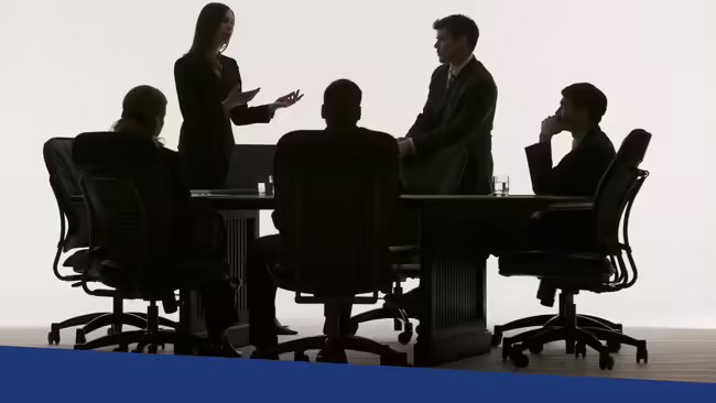 A group of people sitting and standing around a large table having a discussion.