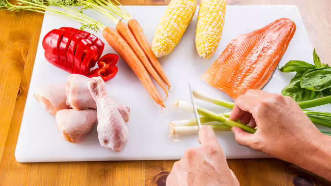 raw meats and vegetables on a cutting board; knife in hand chopping onions