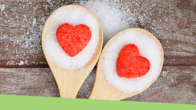 Two wooden spoons sitting on a wooden table. The spoons are full of white sugar with a red heart in the middle of each spoon.
