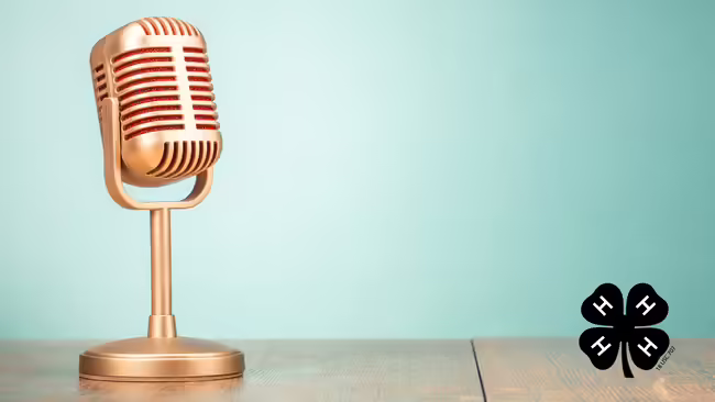 A gold microphone sitting on a wooden desk with a blue background. A black 4-H clover in the bottom right corner.