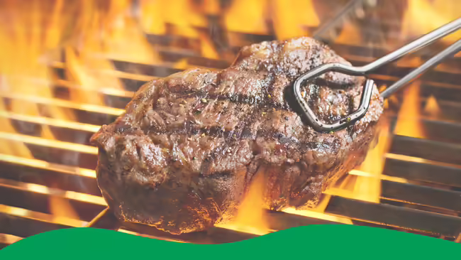 A sirloin steak being held with tongs cooking on a grill with flames around the steak.