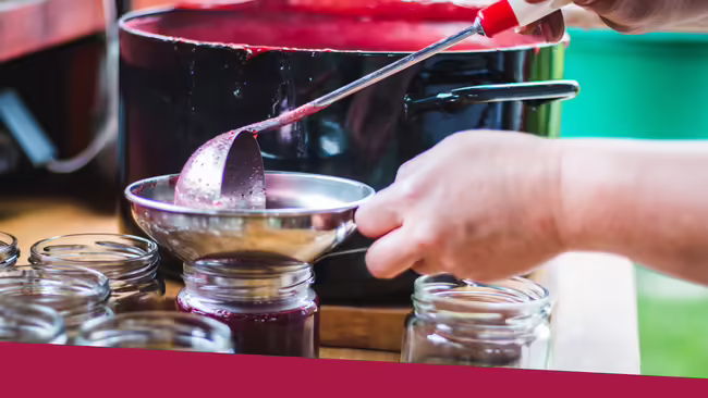 A person holding a funnel inside a glass jar while using a ladle with their other hand pouring red jam into the funnel.