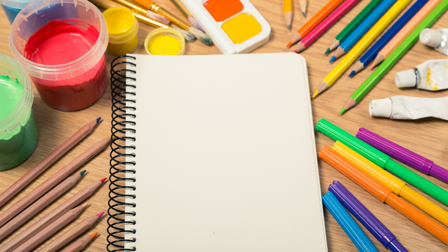 A notebook with many different art supplies surrounds it on a table.