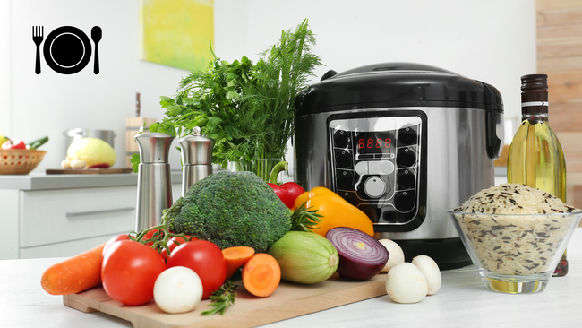 An electric pressure cooker sitting on a kitchen countertop with vegetables, herbs, spices and rice surrounding the cooker.