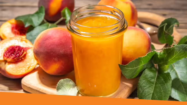 Peach jam in a glass jar with peaches surrounding the jar sitting on a wooden table.
