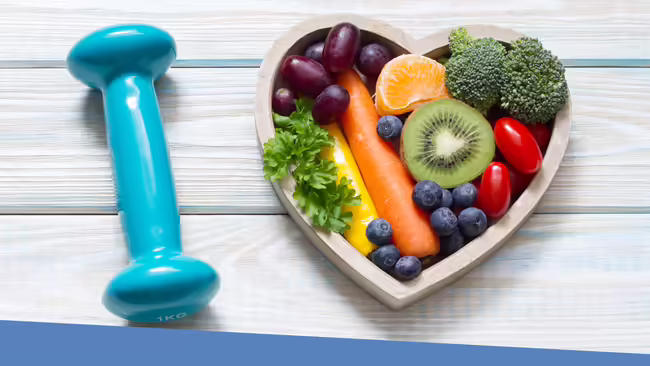 Various fruits a vegetables sitting in a heart shaped bowl, next to the bowl is a small sizes dumbbell.