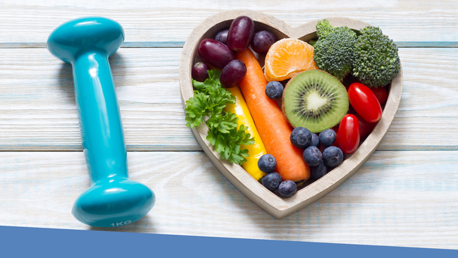 Various fruits a vegetables sitting in a heart shaped bowl, next to the bowl is a small sizes dumbbell. 