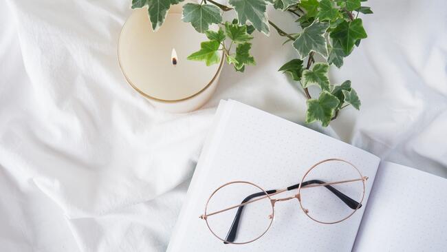 Glasses placed on a writing journal that is next to a lit candle and plant. Gafas colocadas sobre un diario de escritura que está junto a una vela encendida y una planta.