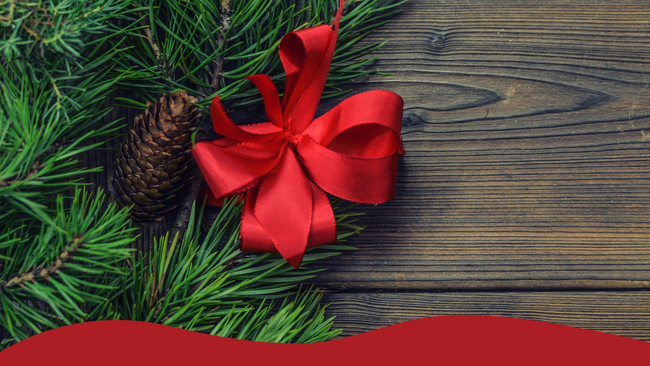 Pieces of evergreen with pinecones and a red bow laying on a wooden table.