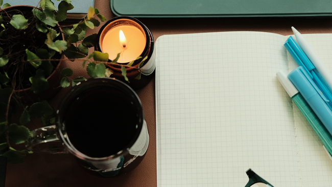 A cup, candle, plant that is next to journal and pens.