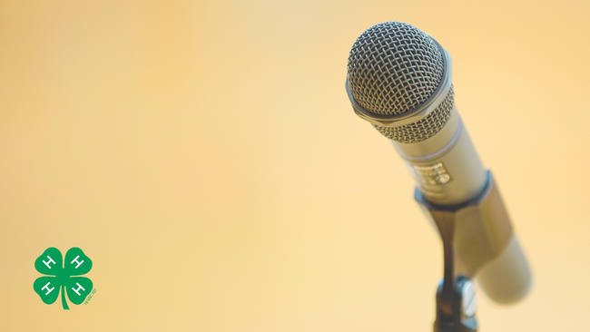 A microphone on a microphone stand on a yellow background with a green 4-H clover in the bottom left corner.