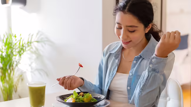 A young woman eating a salad and juice at a kitchen table.