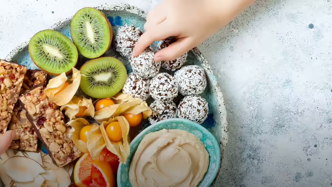 A light blue speckled plate filled with a variety of healthy snacks, with a child's hand reaching in to pick up one of snacks.