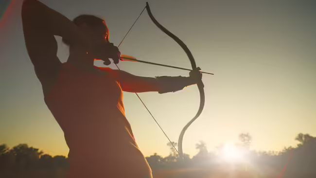 female archer shooting with a bow in a field at sunset