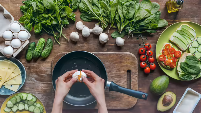 A person cracking an egg into a frying pan, surrounded by various ingredients to make a sandwich.