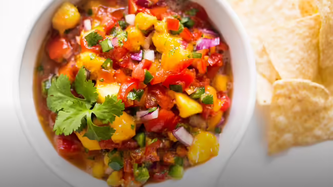 A white bowl with peach salsa in it with tortilla chips next to the side of the bowl.