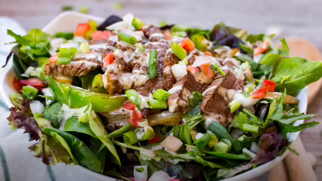 A steak salad in a white bowl, garnished with chopped green onions and diced tomatoes, and drizzled with a creamy white dressing. The bowl sits on a white background with a wooden surface.