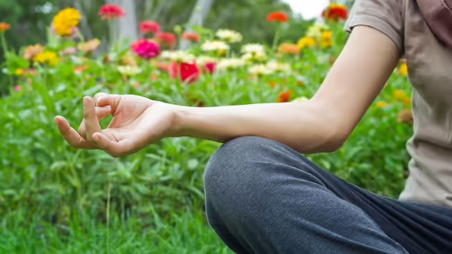 A person sits near a flower garden, with her legs crossed and arm outstretched for a calm yoga pose.
