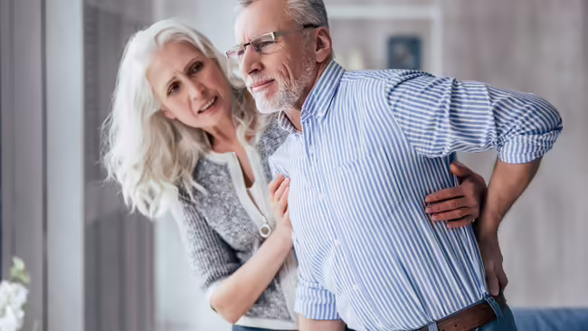 An older couple, the man is grasping his back in pain while the woman supports and looks on with concern.