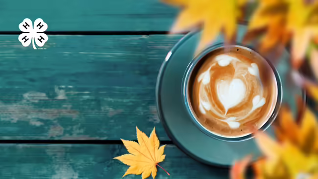 A cup of a latte on a teal wooden table with orange and yellows leaves around the cup. In the top left corner is a white 4-H logo.