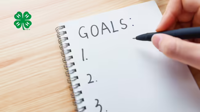 A hand holding a pen over a notebook with the word "GOALS" written at the top. The notebook is on a wooden desk. A green 4-H logo is in the top left corner.