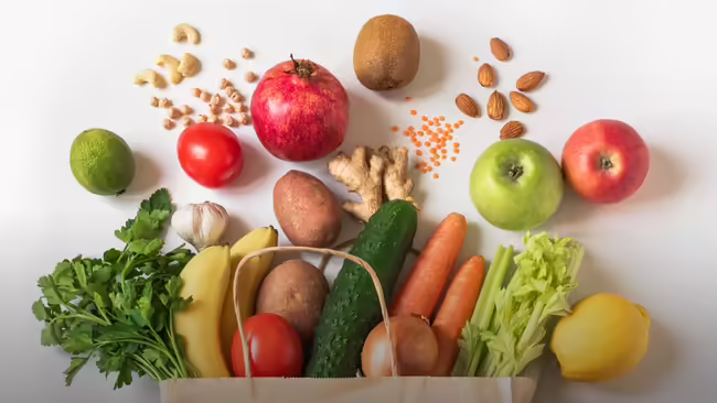 A variety of fruits, vegetables, nuts arranged on a white background, spilling out of a paper bag.