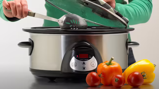 A person lifts the lid on a slow cooker, holding a spoon. In front of the cooker are red and yellow peppers and tomatoes.