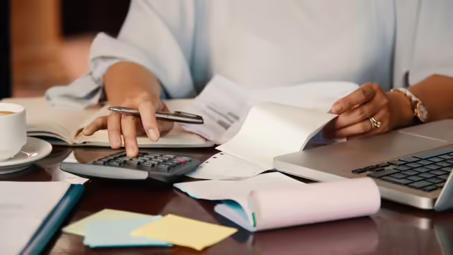 lady using a calculator with laptop and papers on the table all around her