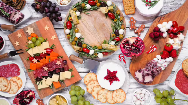 A festive holiday charcuterie spread displayed on wooden boards arranged in shapes such as a Christmas tree and a wreath, set on a rustic white wooden table.