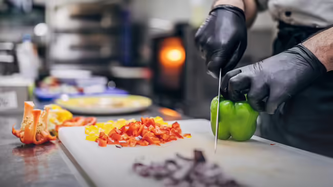 A chef cutting a green pepper on a cutting board with a variety of vegetables that are cut surrounding the cutting board.