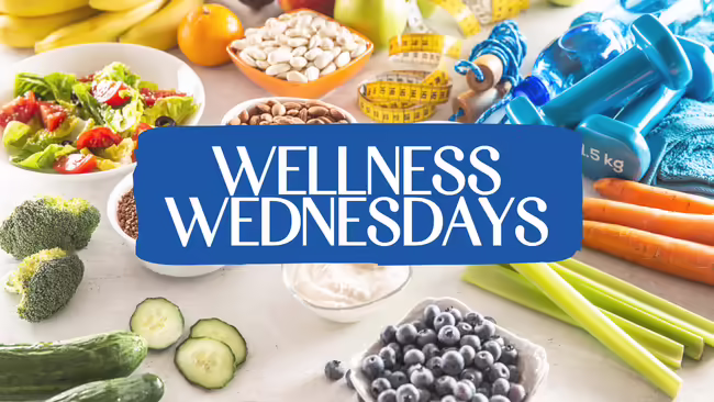 A variety of healthy foods and wellness items displayed on a table, with a blue banner in the center that reads “Wellness Wednesdays.”