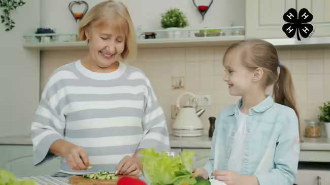 A grandmother and granddaughter in a kitchen. The grandmother chops vegetables on a cutting board. The black 4-H logo is in the upper right corner.