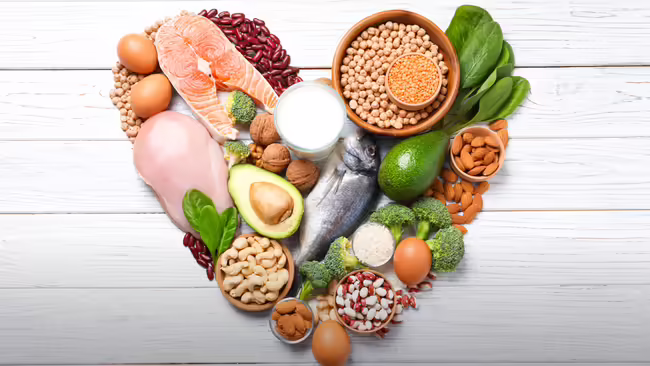 A heart shape made of foods including fish, chicken, eggs, nuts, beans, lentils, avocado, broccoli, and spinach.