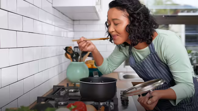 A woman in apron tastes food from a wooden spoon in a kitchen.