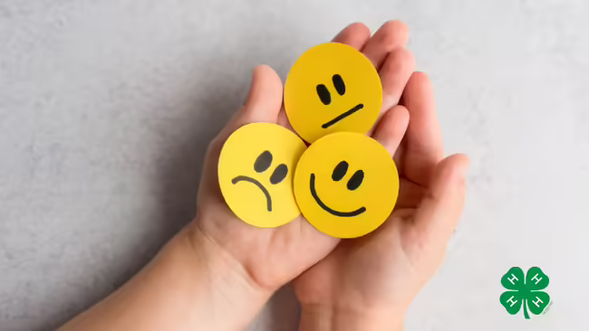 Hands holding paper with hand-drawn expressions: a sad face, a neutral face, and a happy face. A green 4-H clover logo is in the bottom right corner.
