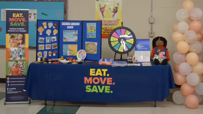 A table covered with a blue tablecloth featuring the Eat. Move. Save. logo, displaying games and informational flyers, with balloons positioned nearby.