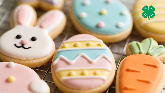 Iced sugar cookies in the shapes of a bunny, eggs, and a carrot on a wire cooling rack with a green 4-H logo in right upper corner.