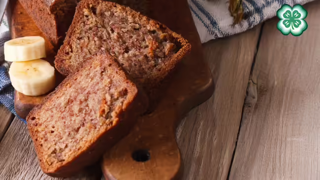 Slices of banana bread on a wooden board with fresh banana slices. A green 4-H clover in the upper right corner.