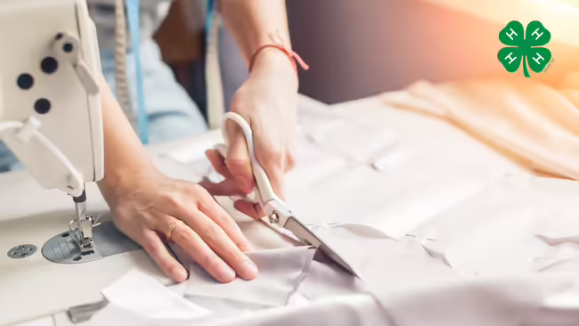 A person cutting fabric next to a sewing machine. A green 4-H clover in the upper right corner.