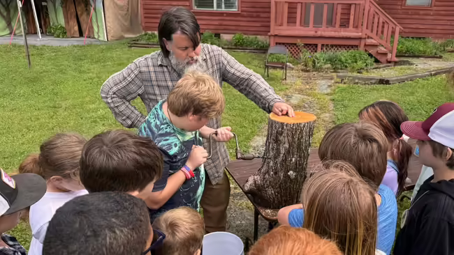 Students gather around a log for a chance to use a hand-tool used for maple syrup production.