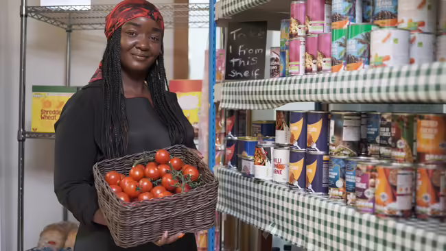 A woman with a box of tomatoes stands in front of a stocked food pantry.