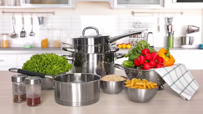 a kitchen in the background and a counter with cooking pots, strainer with vegetables, bowls with lettuce, rice, and pasta, and seasonings nearby