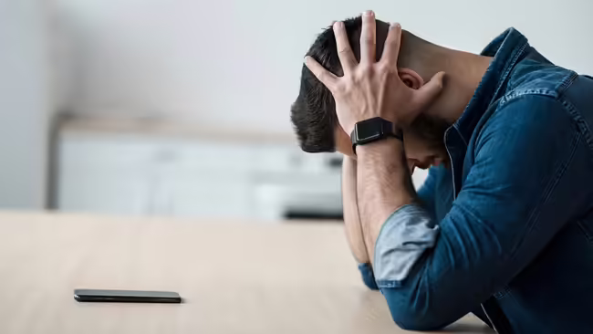 Man in a denim shirt sits at a table with his head rested in his hands.
