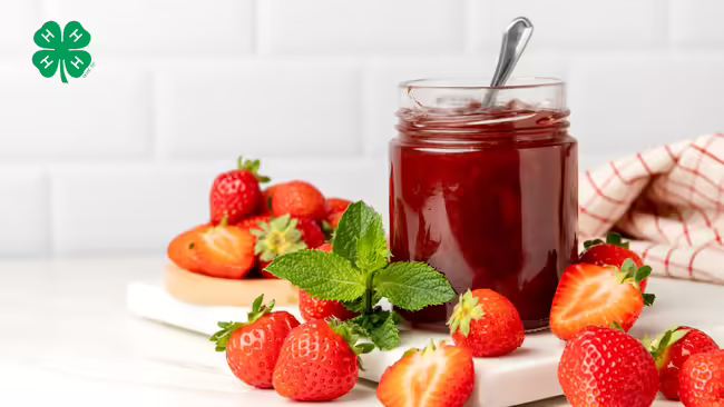 An open jar of strawberry jam with a spoon inside, surrounded by fresh whole and halved strawberries. A green 4-H logo in the top left corner.