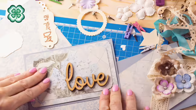 A person arranges a scrapbook page surrounded by craft supplies like lace, paper flowers, and ribbons. A green 4-H clover in the upper left corner.
