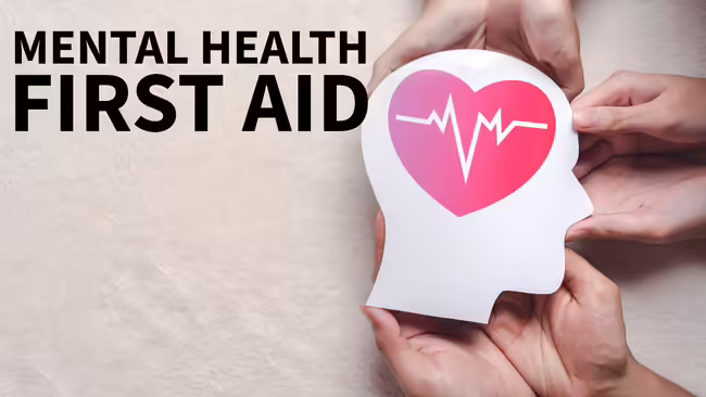 Text reads "Mental Health First Aid". On the right, several hands hold a white paper cutout of a human head silhouette featuring a pink heart.