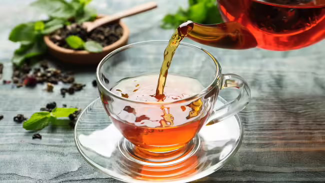  A glass teapot pouring tea into a clear glass teacup on a rustic wooden table with loose tea leaves and mint sprigs in the background.