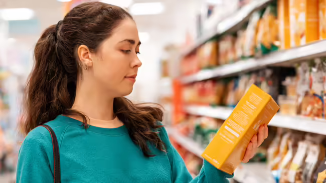 A woman stands in a grocery store aisle, carefully reading the label on a box of food.